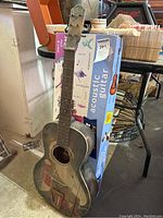 Vintage wooden acoustic guitar leaning against beginner guitar box, showing worn painted design on front with red and beige figures.
