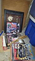 Photo showing framed NASCAR memorabilia including a plaque with number 88 car and other framed photos, Dale Earnhardt themed clock wall plaque, two wooden baseball bats, and some magazines and ticket stubs on floor.