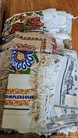 Stack of various Polish linens including a colorful fruit print tea towel, a beige table runner with red traditional embroidery pattern, and a white fringed linen with printed design and Polish text.