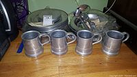 Four pewter mugs in front with various pewter plates and serving utensils behind, displayed on hardwood floor.