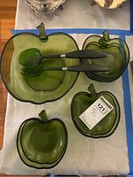 Full view of entire vintage apple-shaped glass salad bowl set including one large bowl, six smaller bowls, and two salad servers, displayed on a table.
