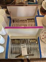 Two boxed sets of dessert flatware showing spoons, forks, and pie servers inside fabric-lined boxes on a table with other dishware items.