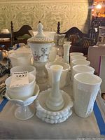 Photo showing a variety of milk glass items on a table including grape pattern pedestal bowls, a lidded pedestal bowl with floral painting, several vases, and smooth round glasses arranged for display.