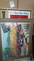 Two framed vintage beer signs leaning against a wall, showing overall lot contents including the Pabst Blue Ribbon baseball bat sign and Miller High Life Indy 500 sign.