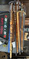 Top view of six carved walking sticks/canes with detailed carved handles and decorative sword with ornate hilt and scabbard, laid side by side on glass table.