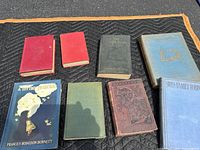 Group photo of eight antique books on a black quilted surface outdoors. Includes varied bindings and colors as described. Books arranged in two rows.