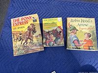 Three children's hardcover books with illustrated dust jackets laid flat showing front covers: The Pony Express, Pocahontas and Captain John Smith, Robin Hood's Arrow.