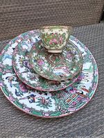 Top view of the stacked dishes and cup showing detailed hand painted designs with floral, people, and animal motifs and gold trim.