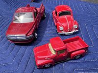 Three red die cast model cars on a blue padded blanket, showing front view of Ram 3500 truck with doors open and top views of the Ford F150 and VW Beetle