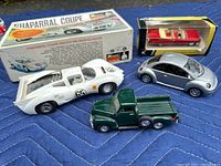 Four die cast model cars displayed on a blue quilted fabric surface: a white Chaparral Coupe racecar model with decals, a silver Volkswagen Beetle, a green six-wheeled Chevrolet pickup truck, and a red Cadillac model inside a clear plastic display box.