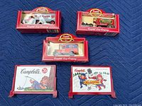 Five die cast model vehicles and billboards from Campbell's Soup collection arranged on a blue surface, showing original packaging and collectible design