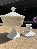 Photo of a white milk glass pedestal dish with lid and a silver crest style candle holder side by side on a table outdoors under sunlight.