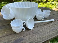 Photo of the midcentury milk glass punch bowl, six matching cups, and plastic ladle shown outdoors on a wooden table.