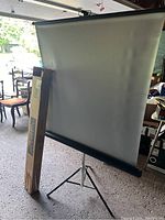 Movie projection screen fully extended on a black tripod stand with original packaging box leaning against it.