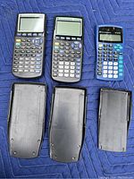 Three Texas Instruments calculators: two dark grey TI-83/83 Plus models and one blue TI-34 II, with backs removed showing battery compartments