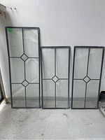 Three antique leaded glass windows standing upright against wall, showing clear geometric diamond pattern design in leaded framework.
