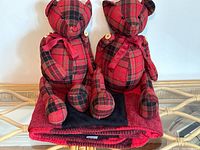 Two black and red plaid wool teddy bears sitting on a folded matching blanket against a white background, showing details of the bears' buttons, scarves, and plaid pattern.