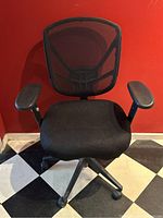 Front view of black mesh office chair with padded seat, armrests, and caster wheel base on checkered floor with red wall background.
