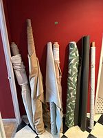 Photo showing eight bolts and rolls of upholstery fabric in various solid colors and leafy print, standing upright against red wall on black and white checkered floor.