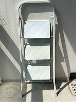 Front view photo of the folded white metal step ladder leaning against a wall, showing the three textured steps.