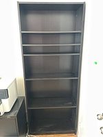Front view of tall black bookshelf with five shelves, showing peg holes for shelf adjustability.