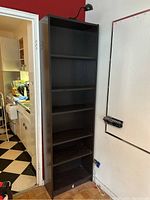 Straight front view of the tall narrow black IKEA bookshelf with six shelves and a black light attached at the top, situated indoors near a kitchen area.
