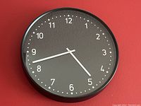 Front view of the black wall clock showing the white numbers, minute dots, and white hands against the black face with a black frame.