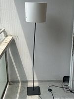 Full view of the tall modern floor lamp standing upright against a white wall with a window to the left, showing the black metal base, slim black stand, and white fabric shade.