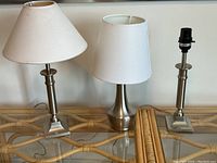 Three modern table lamps on a glass and rattan table, two with white shades and one without a shade, showing chrome bases and shades details.