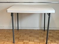 Front view of white plastic folding table with grey metal legs on parquet floor against white wall.