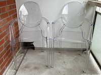 Pair of clear plastic Ghost-style chairs from front, set against brick wall and white wall.