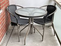 Full patio set on balcony showing round glass top table and two resin wicker chairs with metal frames.