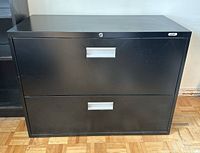 Front view of Staples black 2-drawer filing cabinet on hardwood floor, showing metal handles and lock on top drawer.