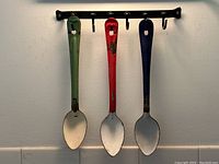 Three vintage large enamel spoons in green, red, and blue, hanging from wrought iron hooks on a wall, showing general wear and paint loss on spoons.