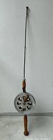 Full view of the vintage fishing rod with metal wire reel against a white wall, showing the full length and wooden handle.