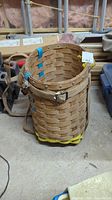 Front angled view of the tall wicker basket backpack with canvas straps, blue tape accents, metal buckle, showing woven wicker construction.