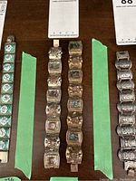 Top-down view of four metal linked bracelets with green tape separators on a wood table. Two bracelets feature abalone shell links.