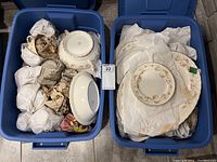 Two large blue plastic bins containing the wrapped china pieces, some plates visible on top in one bin.