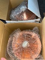 Two copper outdoor table lamps, wrapped in bubble wrap, inside cardboard boxes. Visible lamp tops with copper metal finish.