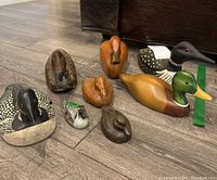 Photo capturing eight wooden bird decoys and loons arranged on floor, showing multiple angles and details including color and wear.