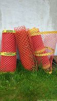 Three rolls of safety orange plastic fencing standing upright against a wall. Each roll is about 48 inches tall, wrapped with yellow caution tape.