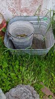 Photo showing one large rectangular galvanized metal wash bin in grass against a wall, containing one galvanized metal bucket inside it.