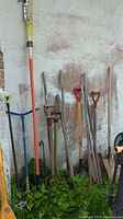 Photo showing the collection of garden tools leaning against an outdoor wall including shovels, pitchforks, hoes, and a garden claw.
