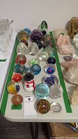 Overview of table with 26 glass orb paperweights arranged in rows, showcasing variety of designs and colors.