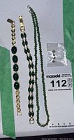 Three jade bracelets, one jade bead necklace, and a pair of small stud earrings with green jade stones on a white surface with MaxSold lot tag.