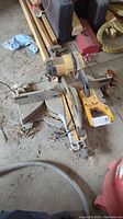 Side angle view of DeWalt miter saw lying on floor showing sliding base, handle and cord with some surrounding items such as wooden planks and cases