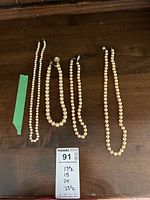 Four knotted pearl necklaces displayed on a wooden surface showing approximate lengths with a MaxSold card listing the sizes.