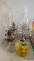 Six glass oil lamps with metal wick holders. Some bases contain colored liquid. Various shapes and sizes with tall glass chimneys.