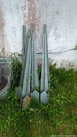 Seven metal post stakes leaning against a wall outdoors on grass, showing overall condition and size.