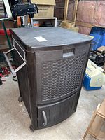 Front angle view of the brown plastic Suncast rolling cooler showing textured front with basket weave design, side metal handle, and wheels.
